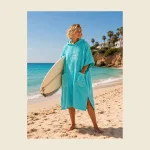Hooded beach poncho in turquoise towelling worn with a surfboard at the shore