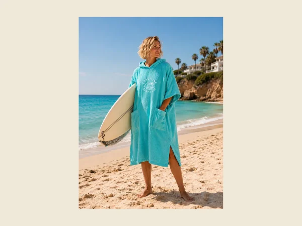 Hooded beach poncho in turquoise towelling worn with a surfboard at the shore