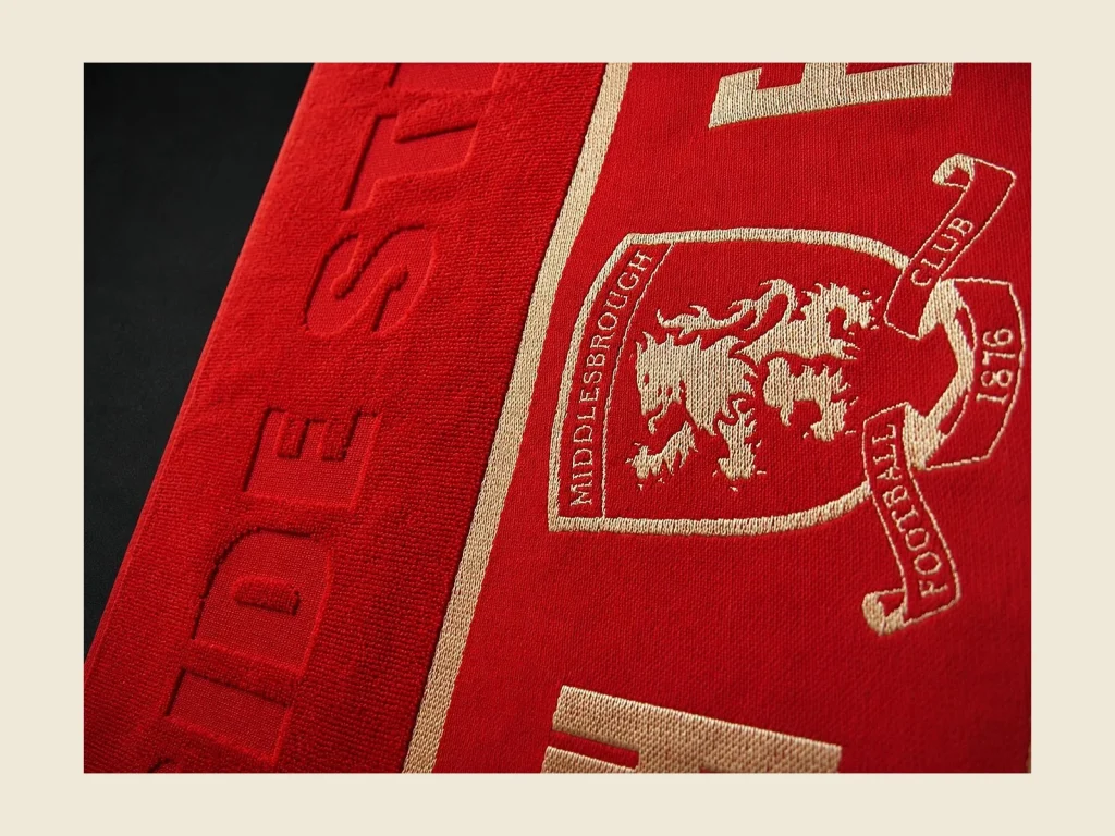 Folded red club towels with embroidered football crests stacked for a sports club programme