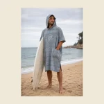Grey hooded surf poncho with a chest pocket worn by a surfer holding a board