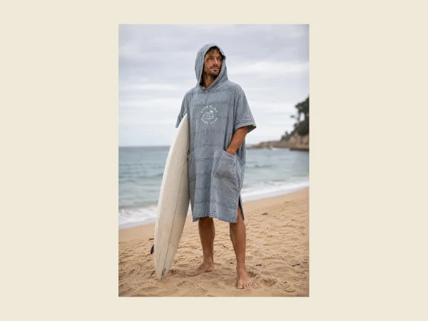 Grey hooded surf poncho with a chest pocket worn by a surfer holding a board