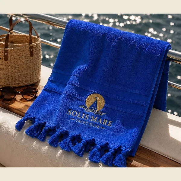 Blue yacht club towel with a gold embroidered emblem and knotted fringe on a boat deck