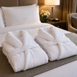 Two folded white terry bathrobes on a hotel bed runner in a guest room with lamp and armchair — MONNEVA