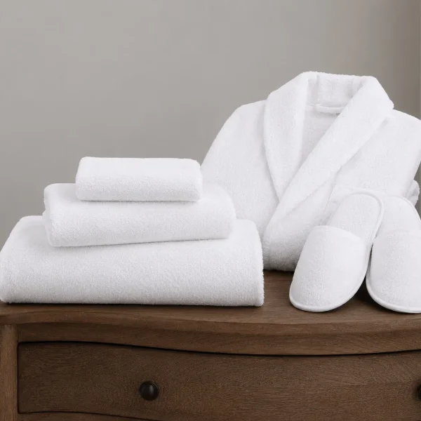 Folded white hotel bath towels with a terry bathrobe and slippers arranged on a wooden dresser