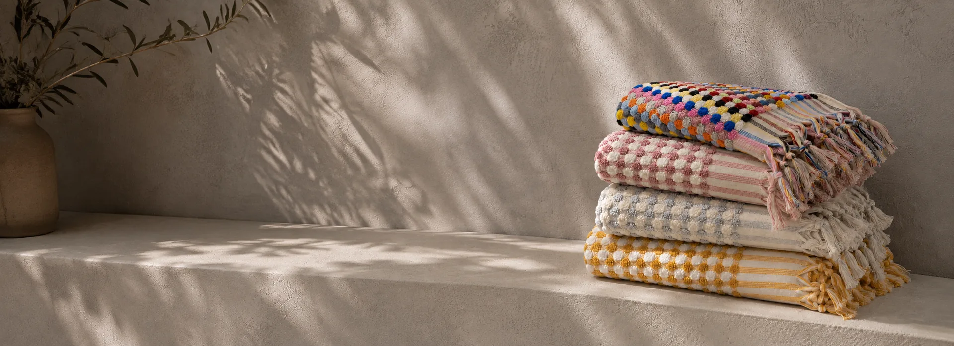 Folded pompom-trimmed peshtemals in multicolour, pink, blue and yellow stacked on a sunlit stone bench — MONNEVA custom colours